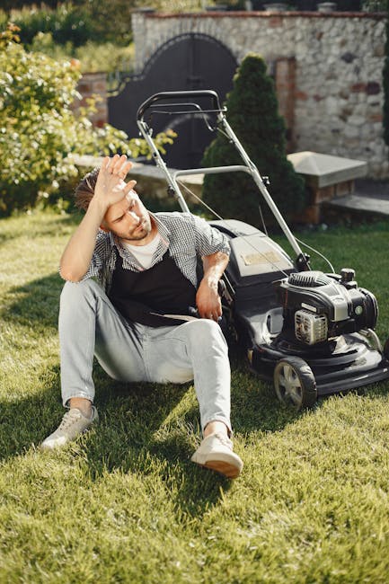 A young man with light brown hair and a beard sitting on a well-maintained, lush green lawn, resting next to a black petrol-driven lawnmower with a silver engine cover. He is wearing a black apron ove